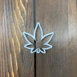 Könnte beinhalten: Ein weißer Kunststoff-Cannabis-Blatt-Ausstecher. Der Ausstecher befindet sich auf einer braunen Holzoberfläche. Der Ausstecher ist ein einzelnes Stück mit einer detaillierten Umrisslinie des Blattes.