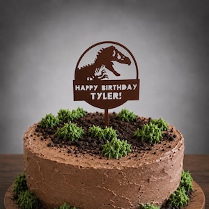 May include: A brown cake topper featuring a dinosaur silhouette inside a circular frame above a rectangular plaque with the text HAPPY BIRTHDAY TYLER! The topper is inserted into a round chocolate frosted cake decorated with green icing and crumbs.