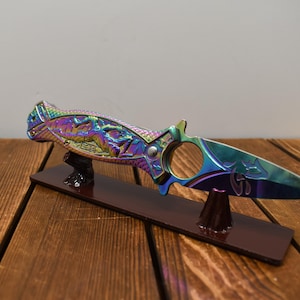 May include: A decorative knife with a rainbow iridescent finish, displayed on a wooden stand. The knife features intricate carvings and a circular opening near the handle. The blade has a pointed tip and is supported by a dark brown wooden base.