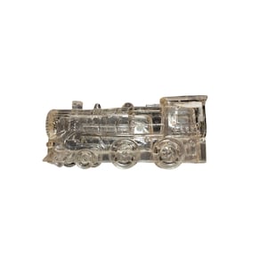 May include: A clear glass train figurine. The locomotive has detailed wheels, a smokestack, and a cab. The transparent design allows light to pass through, creating a shimmering effect. This decorative piece is ideal for collectors or as a unique home accent.