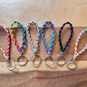 May include: Seven colorful crocheted keychains with silver key rings. Each keychain is a different color gradient, including rainbow, pink, blue, gray, green, black, and orange.