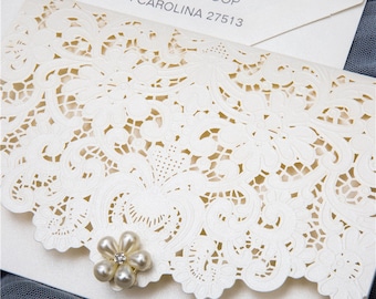 May include: White wedding invitation with a delicate lace pattern and a pearl embellishment. The invitation includes the address "1520 BANYON GROVE LOOP CARY, NORTH CAROLINA 27513".