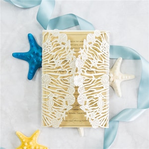 May include: A white laser cut wedding invitation with a seashell and ocean wave design. The invitation is on a beige background with blue and yellow starfish.
