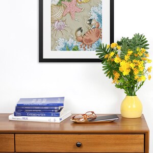 May include: A framed cross-stitch artwork depicting a crab on a sandy beach with seashells and a starfish. The artwork is hanging on a white wall above a wooden console table with books, a cell phone, and a pair of glasses on a gold tray.
