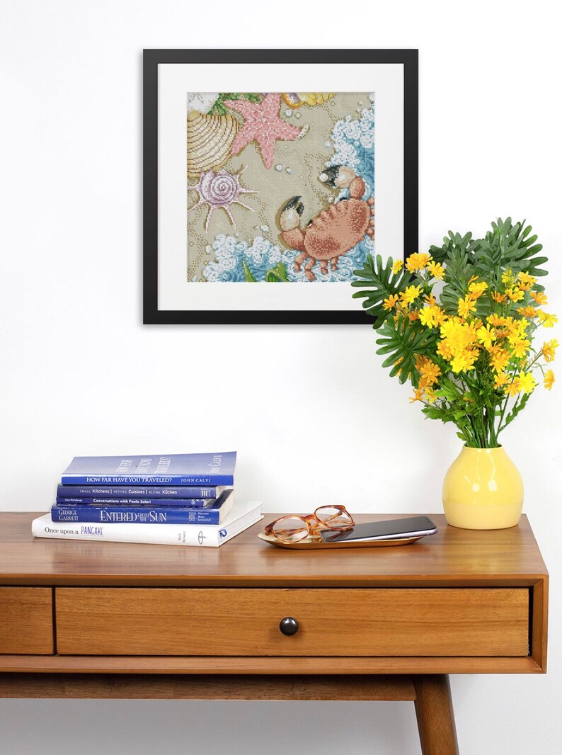 May include: A framed cross-stitch artwork depicting a crab on a sandy beach with seashells and a starfish. The artwork is hanging on a white wall above a wooden console table with books, a cell phone, and a pair of glasses on a gold tray.