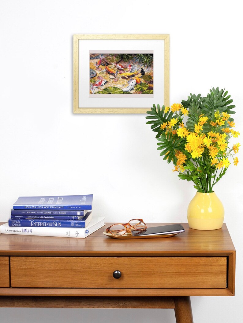 May include: A framed artwork depicting koi fish in a pond, with orange, white, and black markings. The artwork is displayed on a wooden table with books, a tray with glasses, and a yellow vase with yellow flowers.