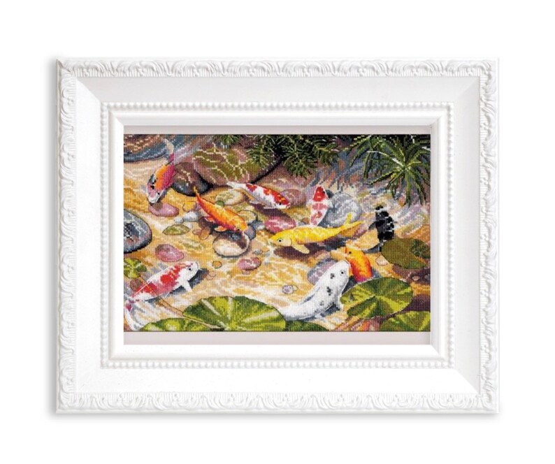 May include: A framed cross-stitch artwork depicts a pond scene with colorful koi fish. The fish are orange, yellow, white, and red, swimming among lily pads and rocks. The frame is white with a decorative edge.