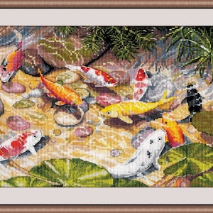 May include: A framed cross-stitch depicting a pond scene with colorful koi fish. The fish are orange, yellow, white, and black, swimming among rocks and lily pads. The water is a light brown color, and the frame is a light brown color.