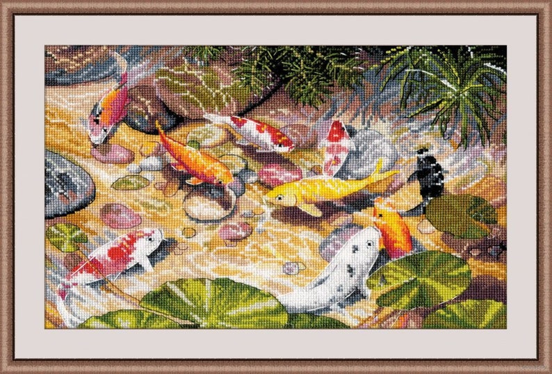 May include: A framed cross-stitch depicting a pond scene with colorful koi fish. The fish are orange, yellow, white, and black, swimming among rocks and lily pads. The water is a light brown color, and the frame is a light brown color.