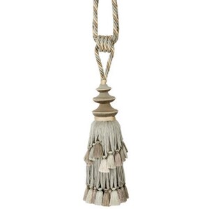 May include: A decorative tassel tieback with a layered, tiered design. The tassel is made of a soft, light gray fabric with a brown and beige trim. The tieback is attached to a rope cord with a gold-colored metal accent.