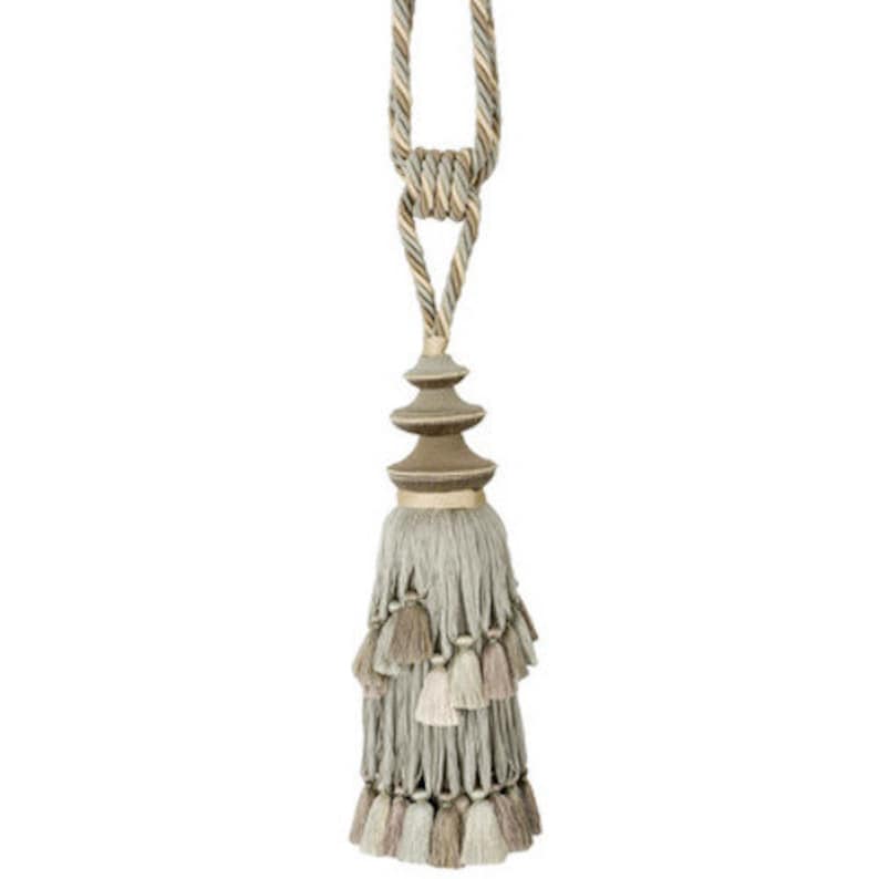 May include: A decorative tassel tieback with a layered, tiered design. The tassel is made of a soft, light gray fabric with a brown and beige trim. The tieback is attached to a rope cord with a gold-colored metal accent.