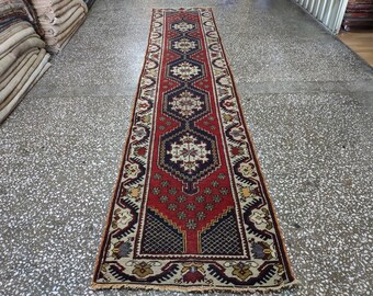 Op de afbeelding: Een lang, smal rood, wit en blauw tapijt met een geometrisch patroon. Het tapijt heeft franjes aan de uiteinden.
