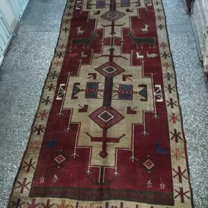 May include: A vintage rug with a red background and a beige center. The rug features a geometric design with stylized animals and a border of repeating patterns.