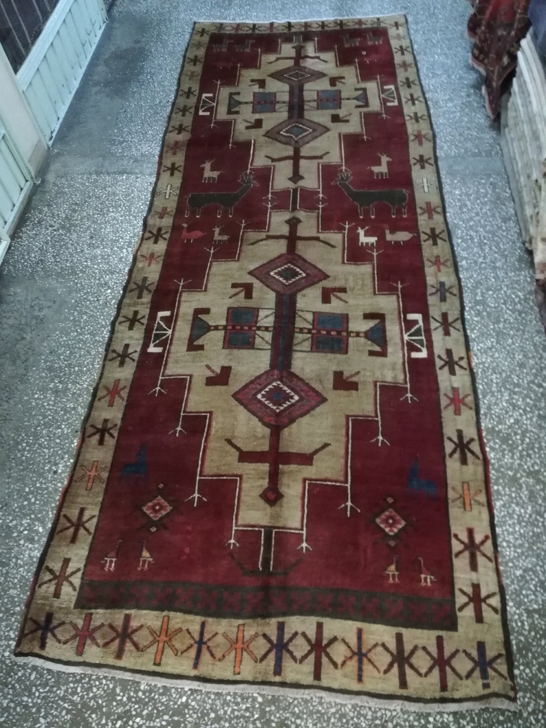 May include: A vintage rug with a red background and a beige center. The rug features a geometric design with stylized animals and a border of repeating patterns.