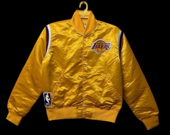 May include: A gold satin Los Angeles Lakers jacket with a button-up front, featuring the Lakers logo on the chest and an NBA logo on the sleeve. The jacket has a ribbed collar, cuffs, and hem, with white and blue accents.