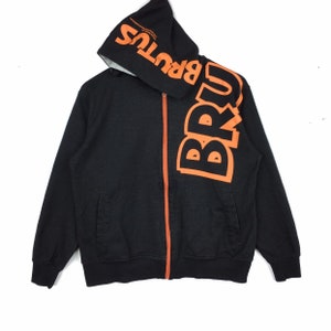 Puede incluir: Sudadera negra con cremallera y capucha con un gran estampado naranja de "BRUTUS" en la parte delantera y la manga.