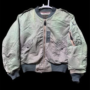 May include: A sage green bomber jacket featuring a ribbed collar, cuffs, and hem. The jacket includes a central zipper closure, two front pockets with snap buttons, a zippered utility pocket on the left sleeve, and shoulder epaulets.