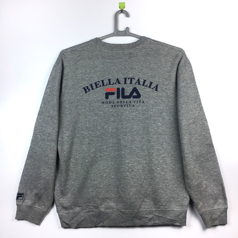Op de afbeelding: Grijs sweatshirt met een Fila-logo en de tekst "Biella Italia" in een gebogen boog boven het logo. De tekst onder het logo luidt "Moda Nella Vita Sportiva".