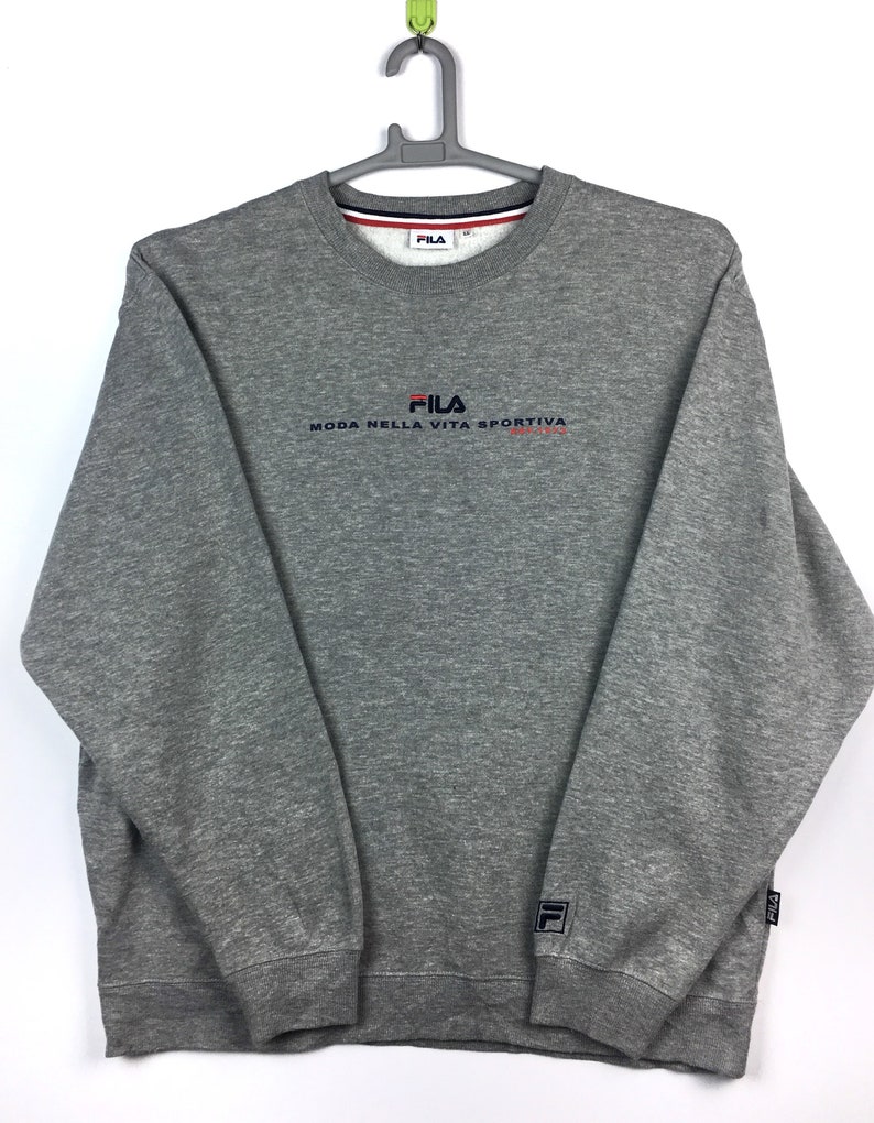 Op de afbeelding: Grijs sweatshirt met een Fila-logo en de tekst "Moda Nella Vita Sportiva" in rood en blauw. Het sweatshirt heeft een ronde hals en lange mouwen.