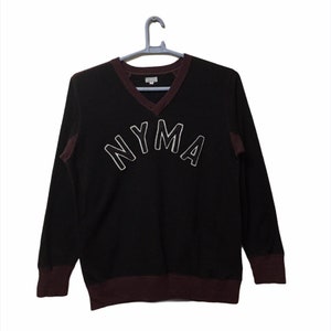 Può includere: Felpa nera a maniche lunghe con scollo a V bordeaux e bordatura bordeaux intorno alle maniche e al fondo. La felpa ha la scritta "NYMA" in lettere bianche bordate sul davanti.