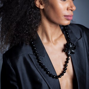 Può includere: Una collana di perline nere con un grande ciondolo a forma di fiore è indossata sopra un blazer nero. La collana presenta perline rotonde e un ciondolo a forma di fiore. Il blazer ha un design classico ed è realizzato in un tessuto lucido.