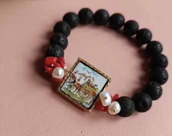 Puede incluir: Pulsera de roca de lava negra con un dije cuadrado con marco dorado que presenta una ilustración colorida de un carruaje tirado por caballos. El dije está acentuado con cuentas de coral rojo y perlas blancas.