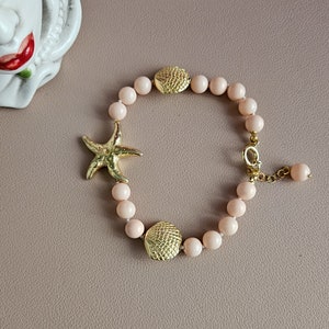 Puede incluir: Un brazalete de cuentas rosadas con abalorios de estrella de mar y conchas doradas. El brazalete está hecho con cuentas pequeñas y redondas y tiene un cierre de langosta.