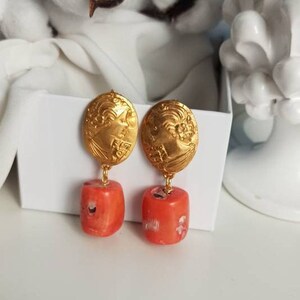 Pu&ograve; includere: Orecchini dorati con un designo dettagliato e un ciondolo di pietra color corallo.