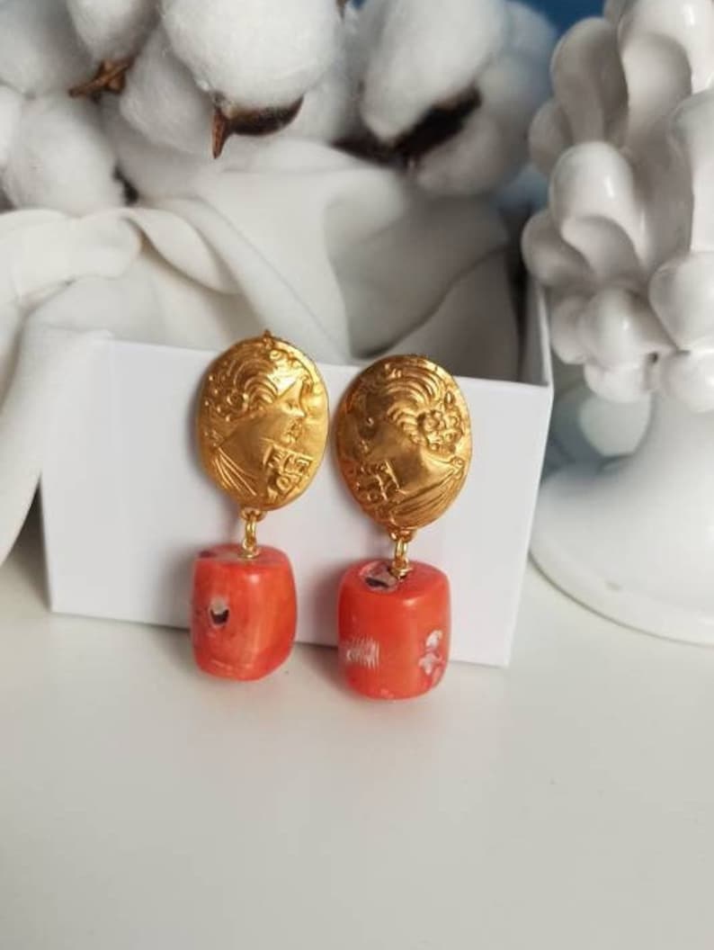Pu&ograve; includere: Orecchini dorati con un designo dettagliato e un ciondolo di pietra color corallo.