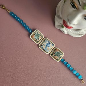 Può includere: Un braccialetto di perline blu con tre ciondoli quadrati raffiguranti creature marine. I ciondoli sono bordati con piccole perline bianche e rifiniture dorate. Il braccialetto ha una chiusura dorata ed è posto su uno sfondo rosa.