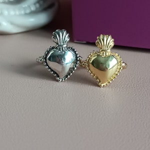Peut inclure: Deux bagues en forme de cœur, une argentée et une dorée, sont présentées. Chaque bague présente un design détaillé avec une pièce maîtresse en forme de cœur et un dessus décoratif. Les bagues sont posées sur un fond rose clair, avec un aperçu d'une boîte violette en arrière-plan.