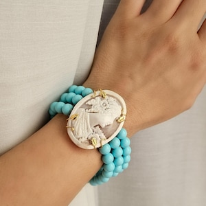 May include: A turquoise blue beaded bracelet with a white cameo featuring a woman's profile. The cameo is accented with gold leaf details.