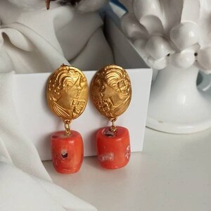 Pu&ograve; includere: Orecchini pendenti dorati con un designo floreale dettagliato e un ciondolo di pietra color corallo.