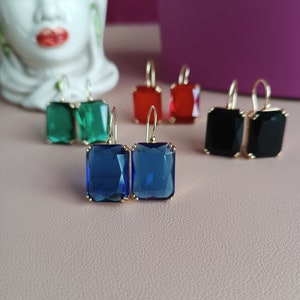 Könnte beinhalten: Ein Set mit sechs Paaren goldener Ohrringe mit quadratischen Glassteinen. Die Steine sind in den Farben Grün, Rot, Blau und Schwarz.