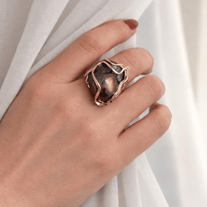 Può includere: Un anello vistoso con una pietra scura e arrotondata incastonata in una montatura color rame a forma di filo. L'anello è indossato su una mano, con un tessuto bianco sullo sfondo. Il design dell'anello è organico e accattivante.