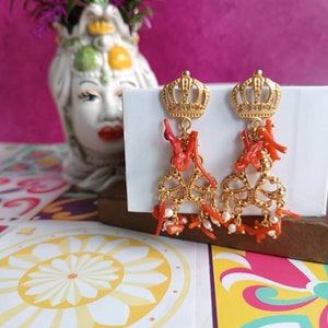 May include: Gold earrings with a crown design and red coral accents. The earrings feature a delicate chain with small white pearls.