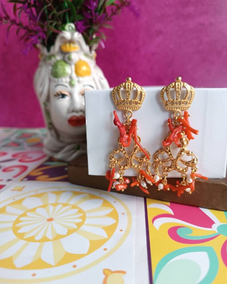 May include: Gold earrings with a crown design and red coral accents. The earrings feature a delicate chain with small white pearls.