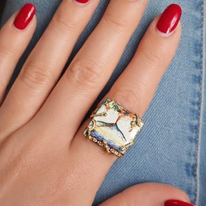 Puede incluir: Un anillo cuadrado con un diseño pintado que presenta una imagen blanca y azul, engastado en un marco de metal dorado. El anillo se lleva en un dedo con esmalte de uñas rojo.