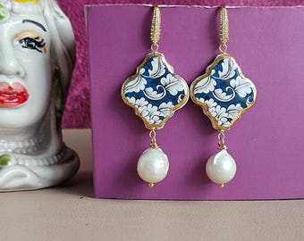 May include: A pair of gold-toned earrings with a navy blue and white floral design. Each earring has a quatrefoil shape with a gold border and a large white pearl drop. The earrings are displayed against a purple background.