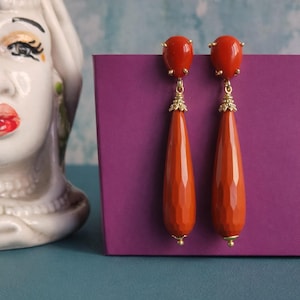 Può includere: Un paio di orecchini pendenti arancioni con dettagli color oro. Gli orecchini hanno un design sfaccettato a goccia e una piccola pietra rotonda arancione nella parte superiore. Gli orecchini sono esposti su una superficie viola.