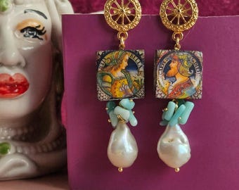 Puede incluir: Pendientes de oro con un diseño de rueda en la parte superior, una baldosa cuadrada con una imagen colorida de una persona, piedras turquesa y una perla blanca grande.