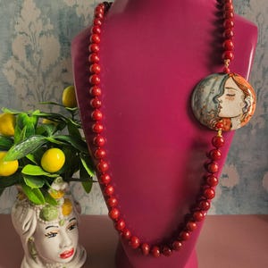 Può includere: Una collana di perline rosse con un ciondolo rotondo raffigurante un profilo dipinto di un volto femminile. La collana è esposta su un manichino magenta. Sullo sfondo un vaso decorativo con limoni e verde.