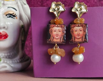 May include: Gold tone earrings with a square portrait of a woman, orange stones, and a pearl drop.