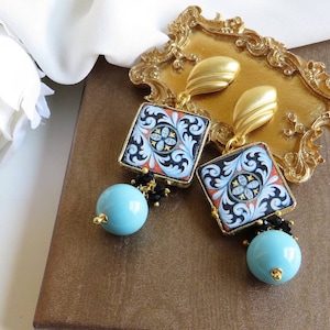 May include: A pair of dangle earrings with blue and gold tile-inspired designs. The earrings feature black beads and a turquoise blue bead at the bottom.