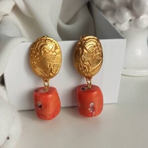 Pu&ograve; includere: Orecchini in tinta oro con un design ovale dettagliato e una perla di corallo pendente.