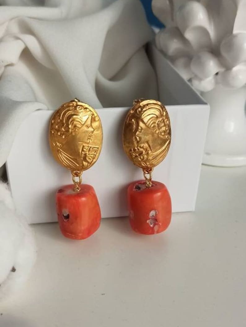 Pu&ograve; includere: Orecchini in tinta oro con un design ovale dettagliato e una perla di corallo pendente.