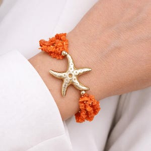 Op de afbeelding: Goudkleurige armband met zeesterhanger en oranje koraalaccenten. De armband heeft een gedetailleerde zeesterhanger en clusters van helder oranje koraal. Een stijlvol accessoire.