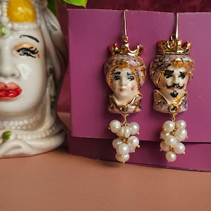 Può includere: Un paio di orecchini dorati con figure in ceramica di un re e una regina che indossano corone. Ogni figura è dipinta con dettagli rosa, bianchi e dorati. Gli orecchini sono adornati da un gruppo di perle bianche.
