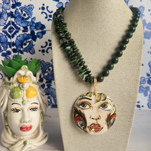Op de afbeelding: Een ketting met een ronde keramische hanger met een handgeschilderd gezicht in wit, goud, rood en zwart. De hanger hangt aan een meerstrengs ketting bestaande uit donkergroene en gespikkelde groen met magenta edelsteenkralen.