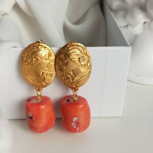 Pu&ograve; includere: Orecchini dorati con un designo floreale dettagliato e perline color corallo che pendono dalla parte inferiore.